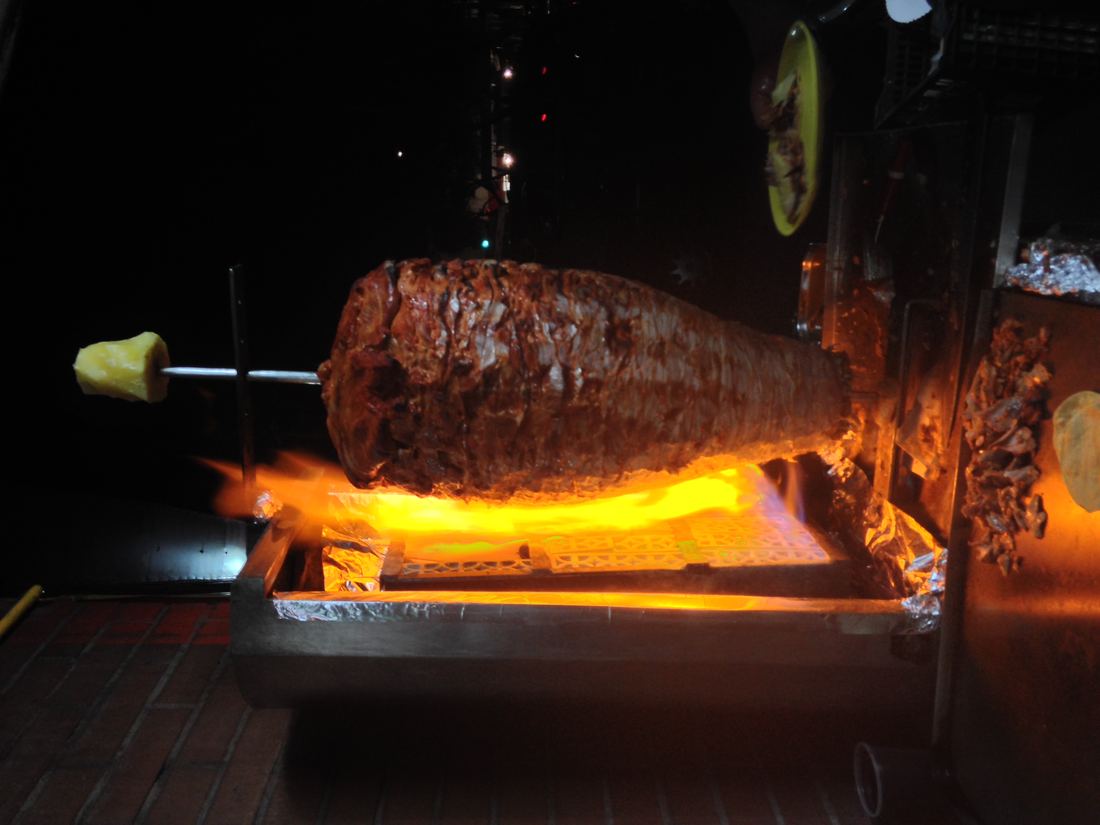 A vertical spit of marinated pork for tacos al pastor glowing beside a flame