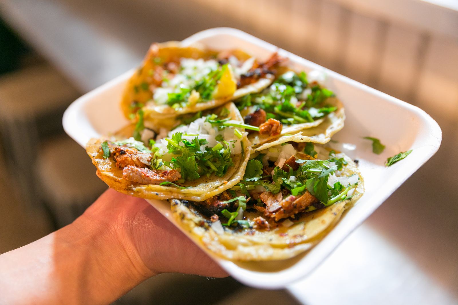 Hand holding a tray of al pastor tacos with onion and cilantro